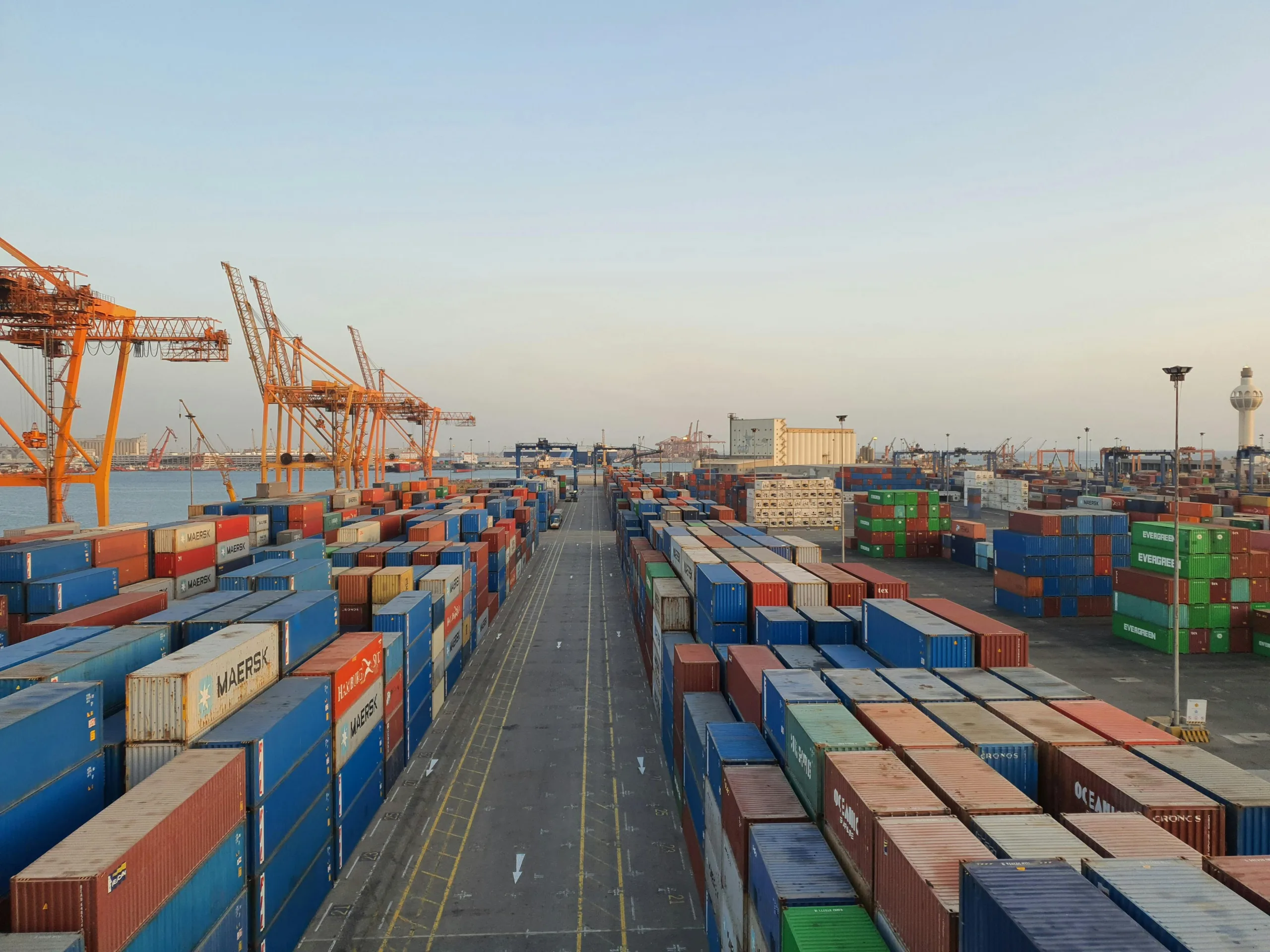 A busy shipping container port with numerous colorful cargo containers stacked in rows, large orange cranes, and a calm sea in the background.