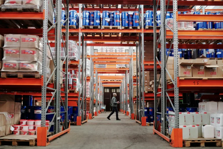 A warehouse with rows of shelves stacked with various products. A person walks between the aisles, observing the items.